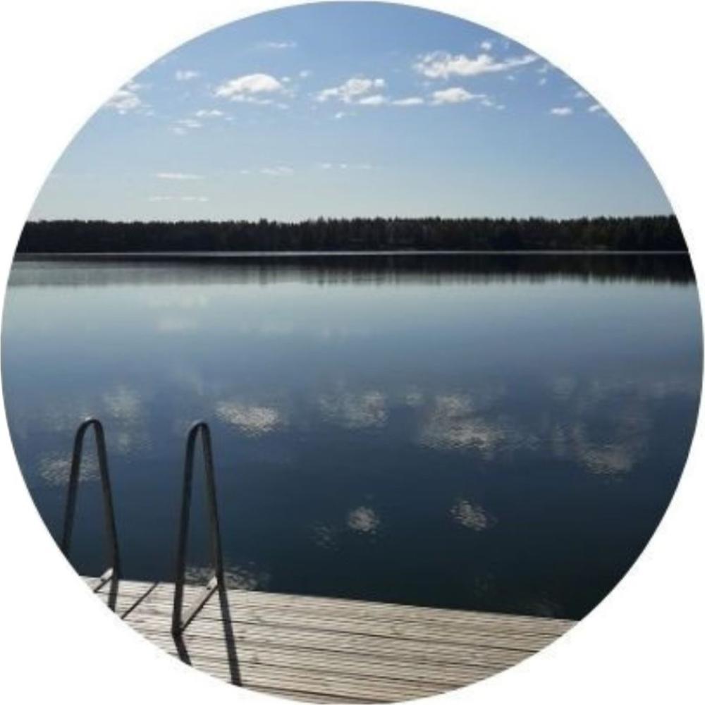 Suomalainen järvimaisema. Sininen taivas, metsä horisontissa ja etualalla laiturin nokka, jossa veteen vievät portaat.