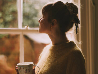 A woman is looking out a window and she is holding a mug in her hand.