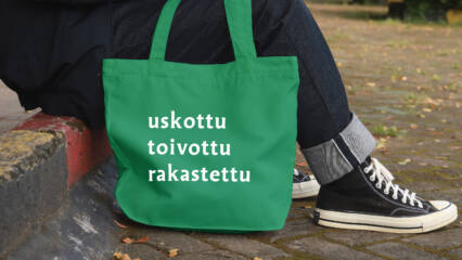 Ihmisen jaloissa kangaskassi, jossa lukee uskottu, toivottu, rakastettu.