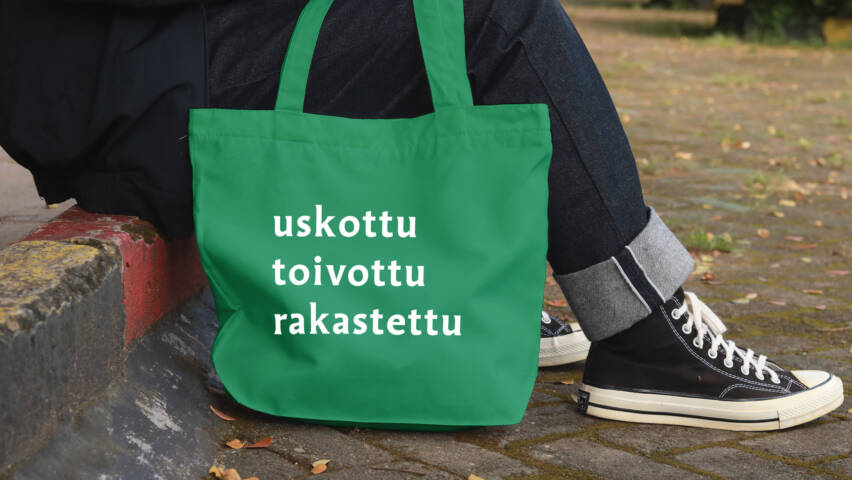 Ihmisen jaloissa kangaskassi, jossa lukee uskottu, toivottu, rakastettu.