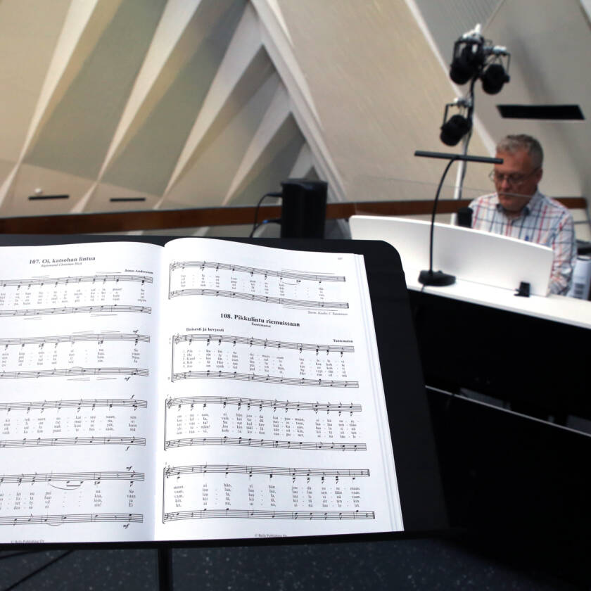 Nuottiteline, jossa kuoronuotit, taustalla mies soittaa pianoa.