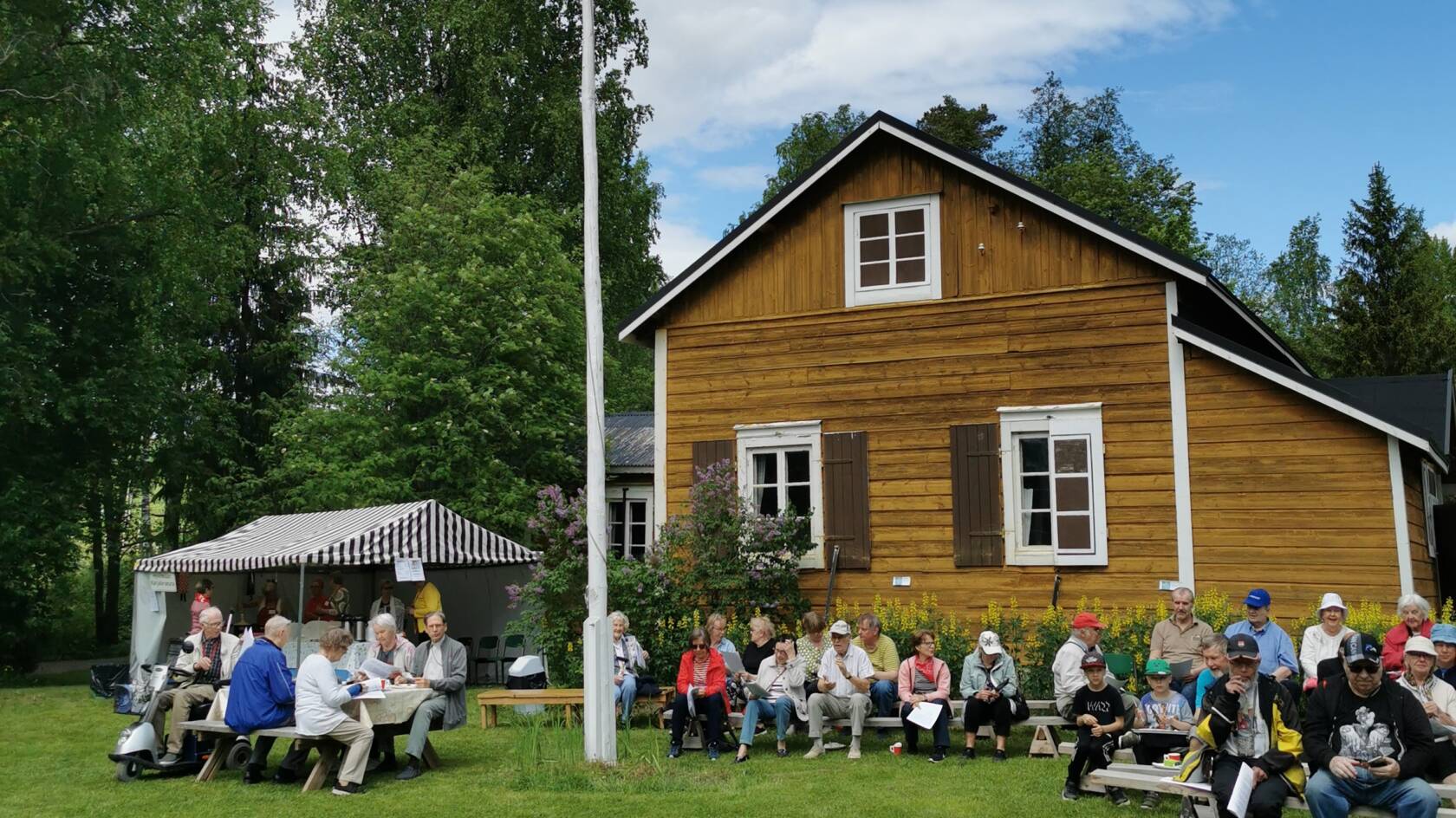 myllytilan keltainen rakennus jonka edessä nurmella ihmisiä viettämässä kesäpäivää istuskellen.