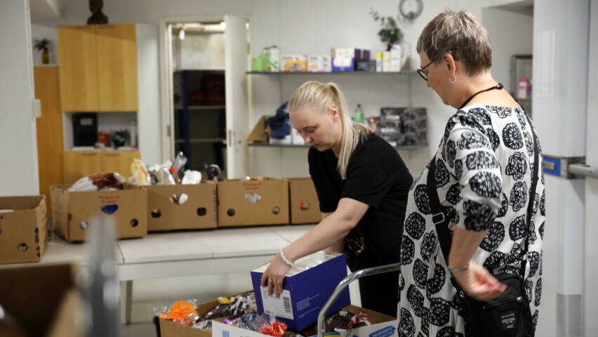 Sara Tiirola ja Maria Kahra lastaavat kärryyn pahvilaatikoita, joissa on ruokaa. Taustalla lisää pahvilaatikoita täynnä ruokaa ja hylly, jossa elintarvikkeita.
