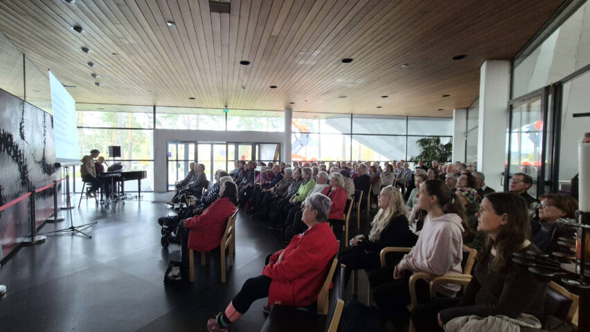 Täydet penkkirivit ihmisiä laulamassa Hyvinkään kirkon aulatilassa.